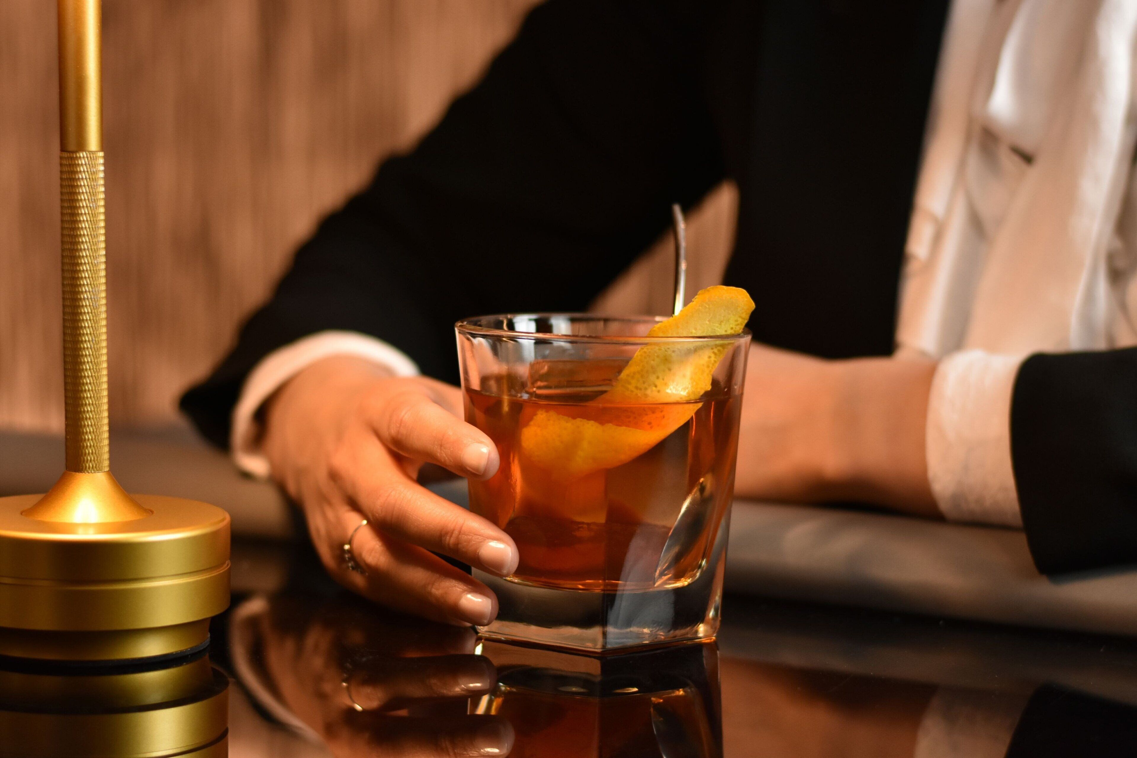 A clear glass with a lemon slice garnish, a gold-colored object beside it, and a person's hand holding the glass.