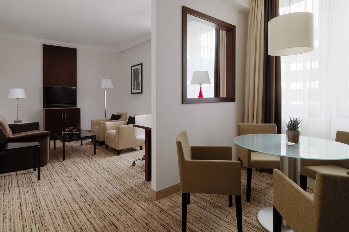 Hotellihuoneessa on lasinen ruokapöytä, beigenväriset nojatuolit, sohva, televisio ja suuri peili.