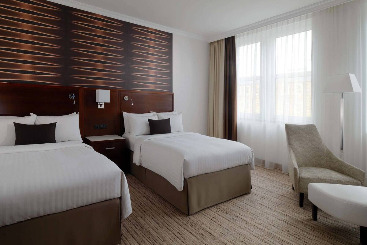 Hotellihuone, jossa on kaksi sänkyä, tuoli, yöpöytä ja suuri ikkuna, jossa on verhot.