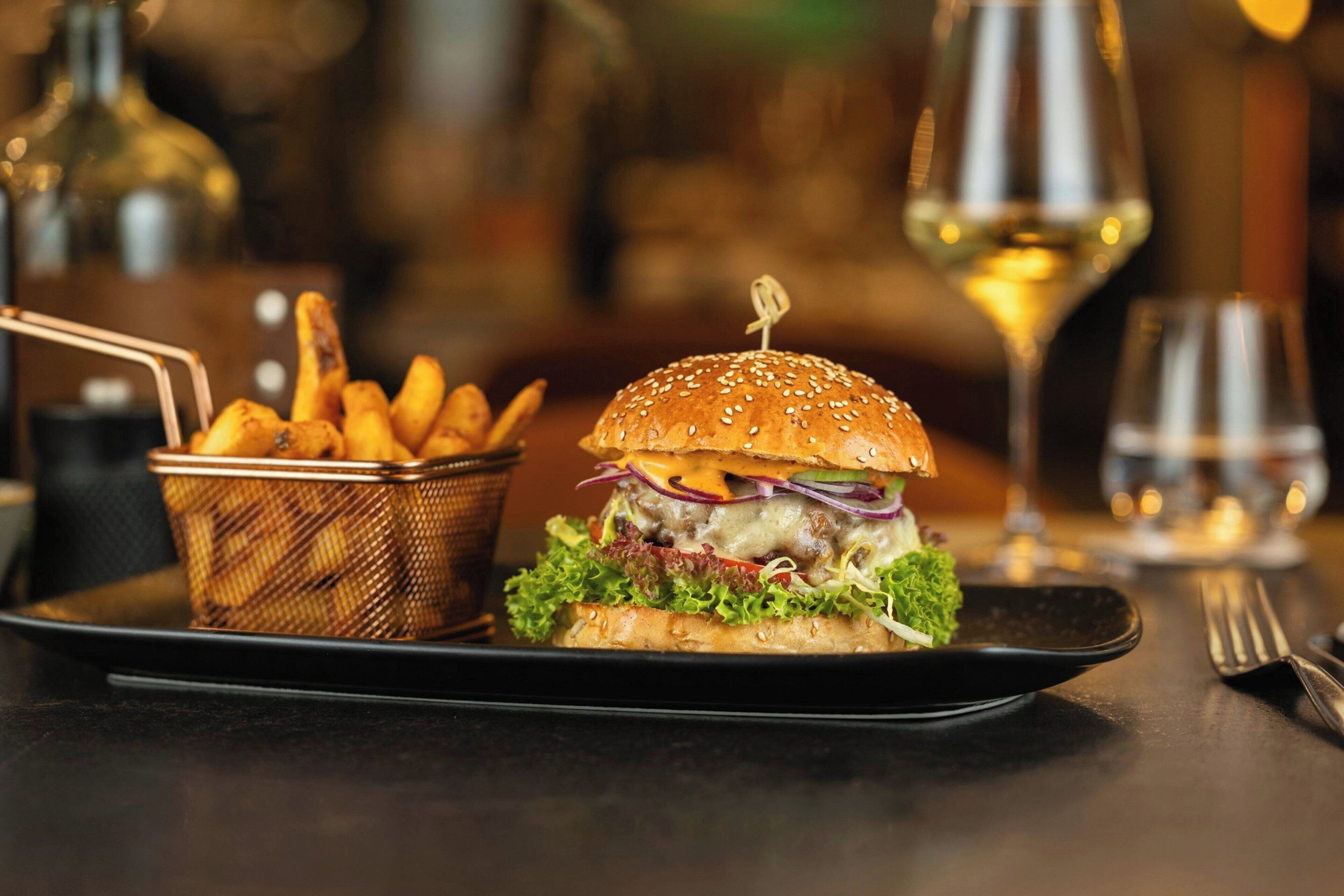 A burger with fries on a black plate, accompanied by a glass of white wine and a wine glass.