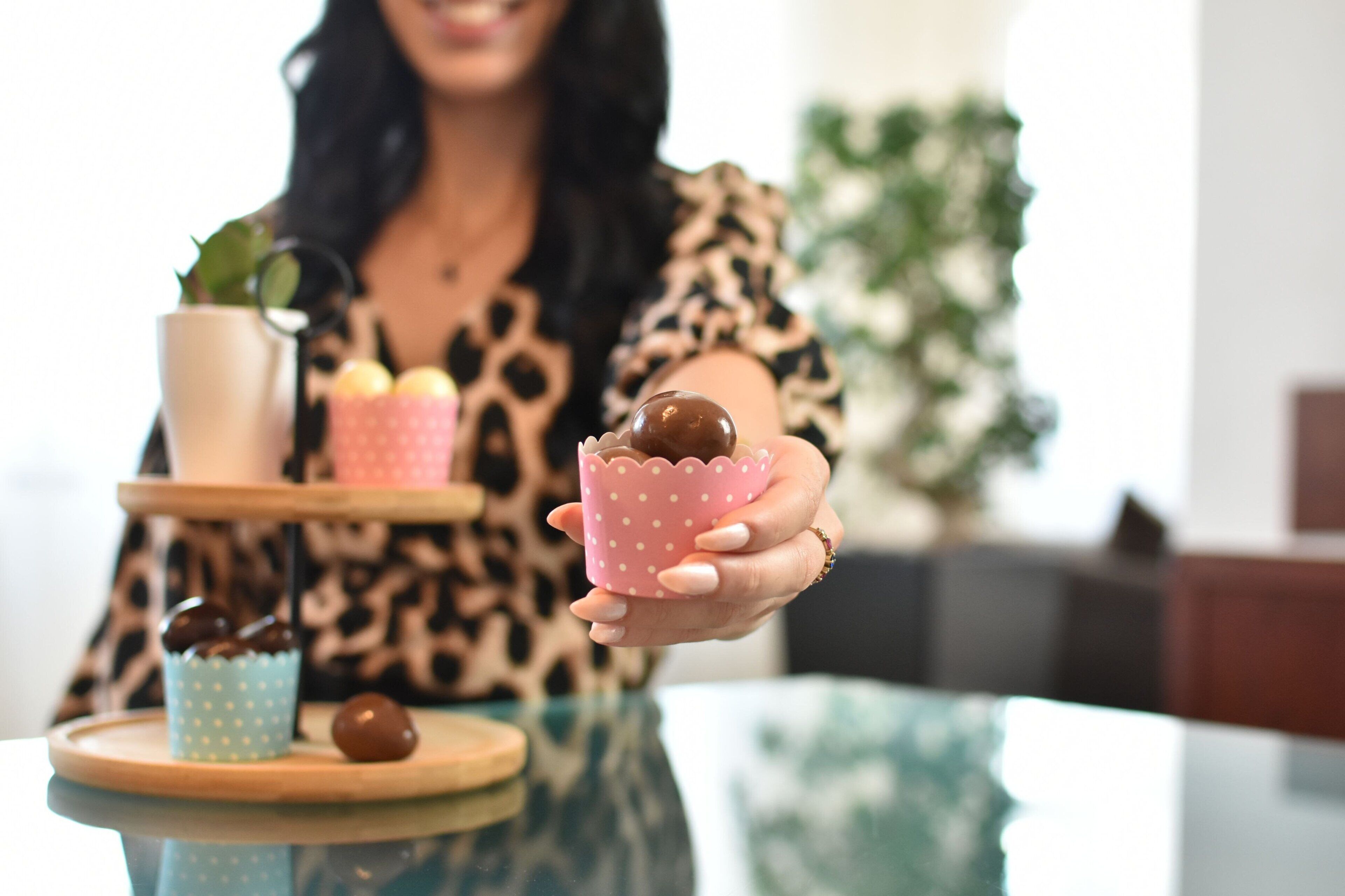 A person holding a pink polka-dotted cup with a chocolate ball, a blue polka-dotted cup, and a white cup with a plant on a wooden tray.