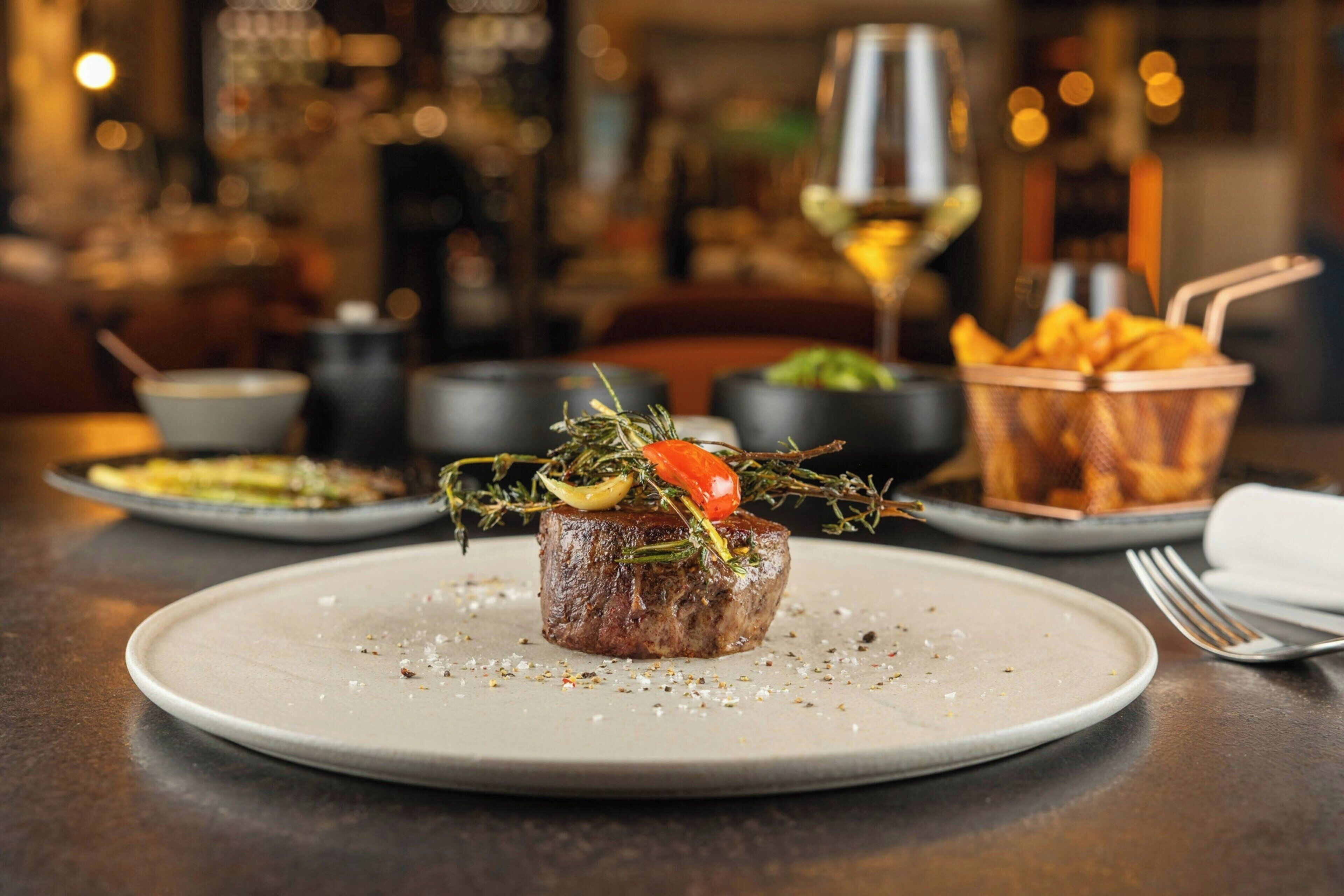 A plated steak with garnish, a glass of wine, and a basket of fries on a table.
