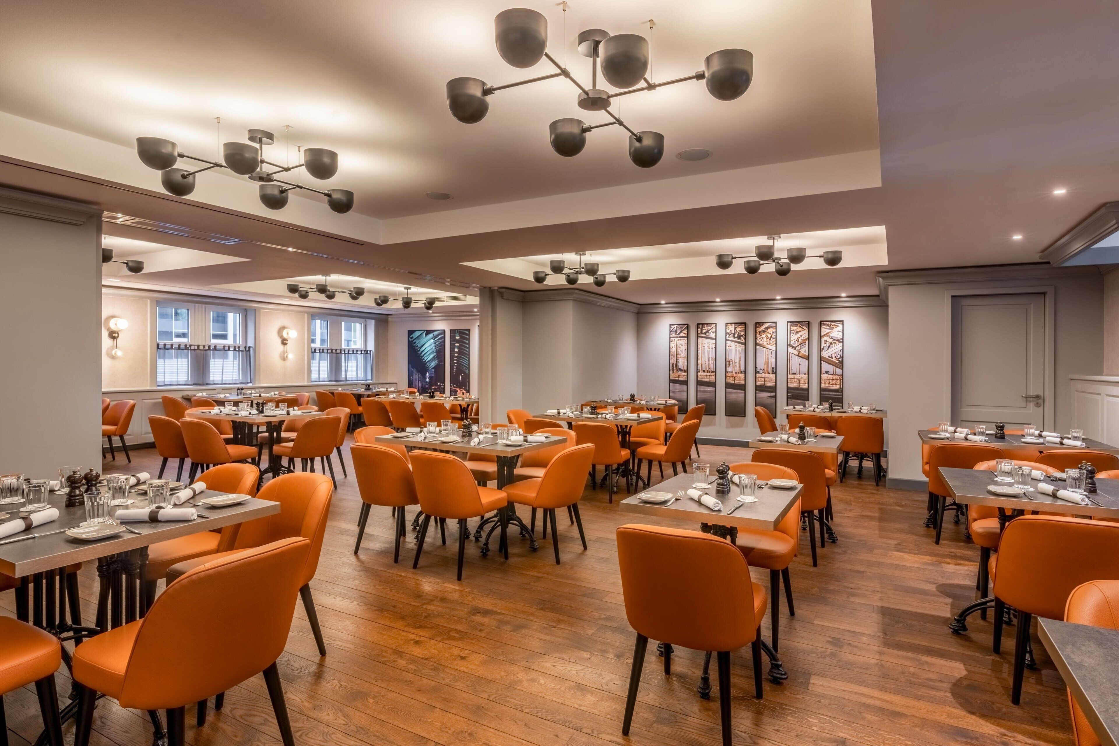 A spacious dining area with multiple tables and orange chairs, featuring modern lighting fixtures and large windows.