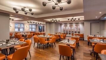 A spacious dining area with multiple tables and orange chairs, featuring modern lighting fixtures and large windows.