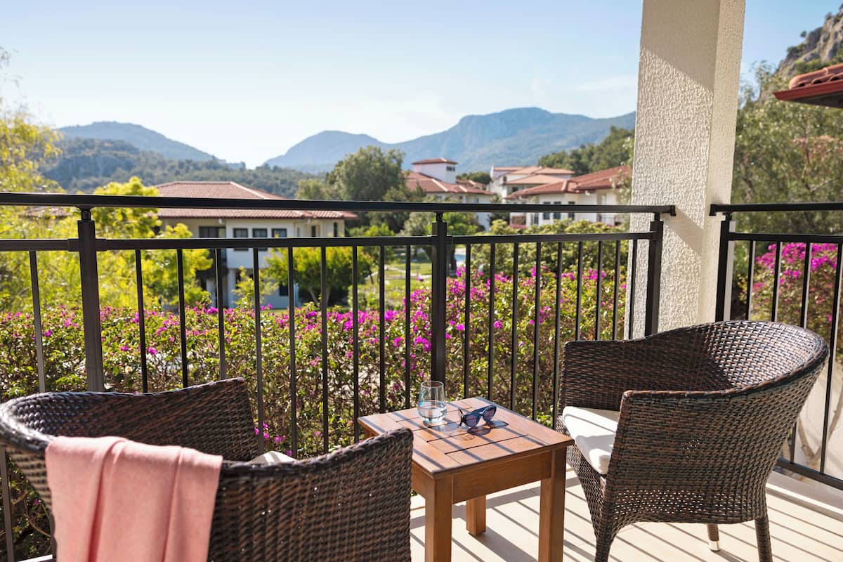 Ein Balkon mit Korbstühlen und einem kleinen Tisch, mit Blick auf einen Garten mit rosa Blumen und Häusern im Hintergrund.