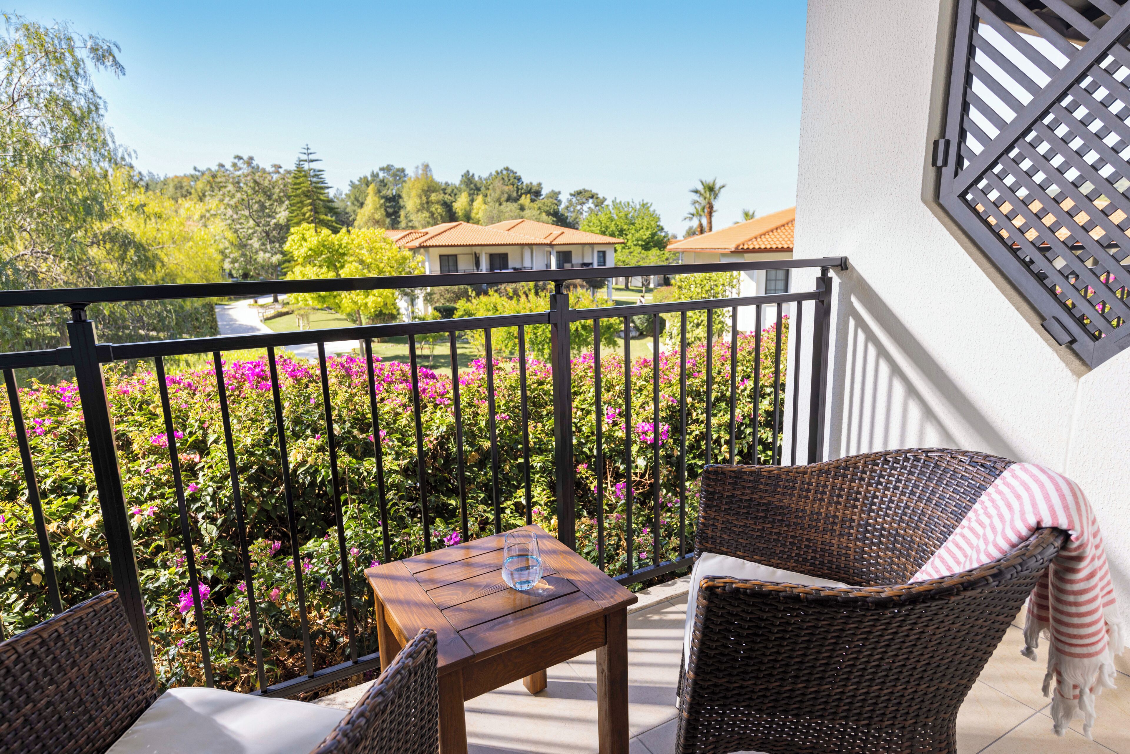 A balcony with wicker furniture, a table, and a view of a garden with pink flowers and a house in the background.
