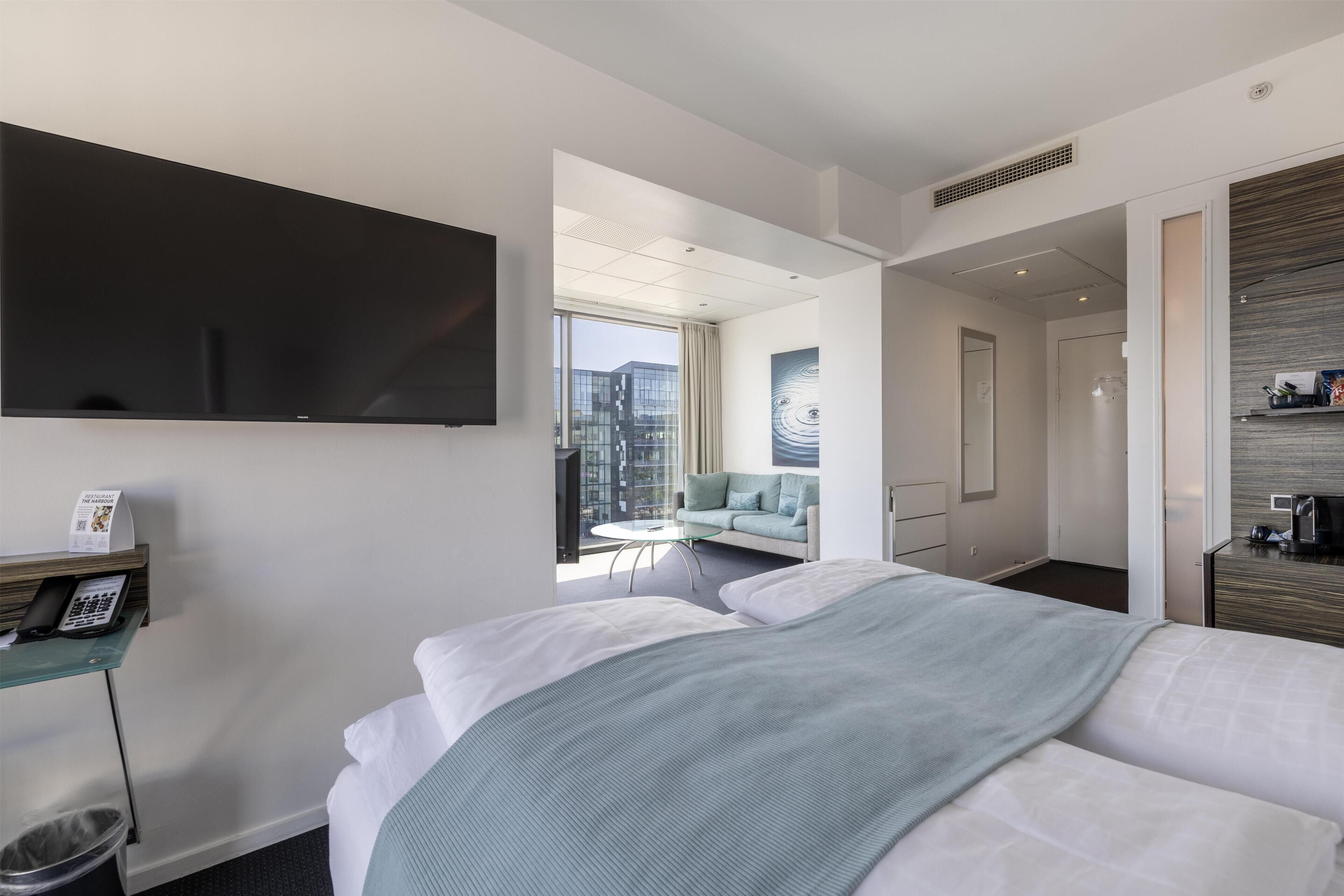 A modern hotel room with a large bed, a flat-screen TV, a desk with a telephone, and a view of the city through large windows.