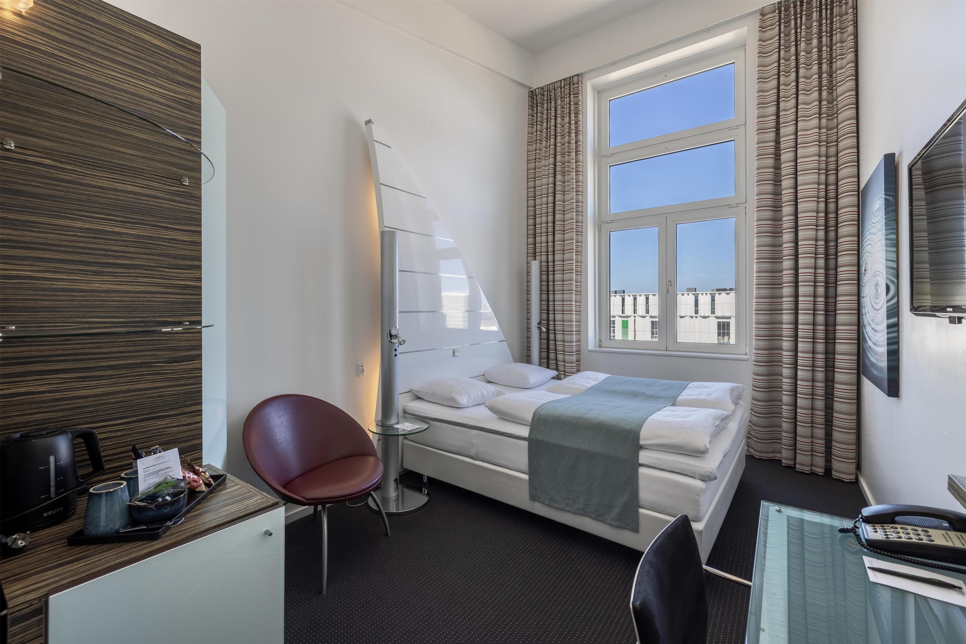 A hotel room with a bed, a desk with a telephone, a chair, a small table with a kettle, and a window with curtains.