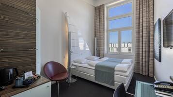 A hotel room with a bed, a desk with a telephone, a chair, a small table with a kettle, and a window with curtains.