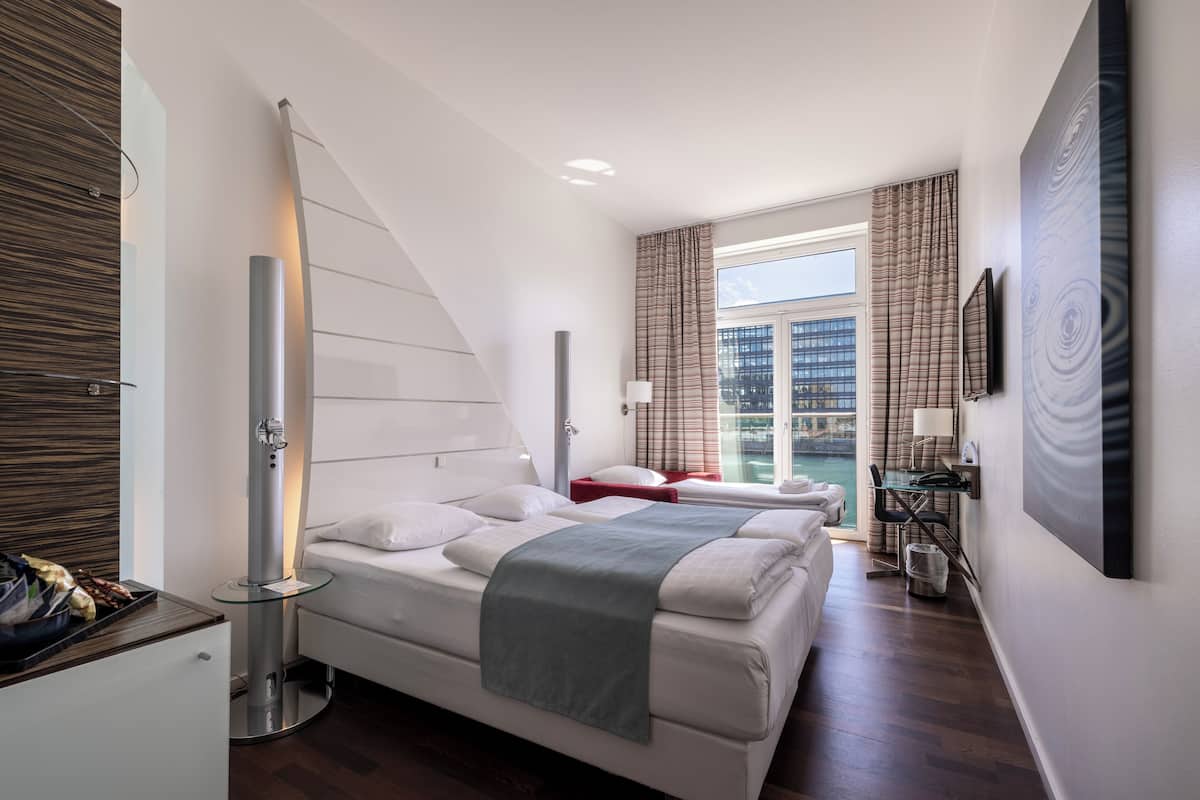 Une chambre d’hôtel moderne avec deux lits, un bureau et une vue sur un immeuble, visible à travers de grandes fenêtres.