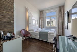 Una habitación de hotel pequeña con una cama, un escritorio con computadora, una silla y un ventanal con cortinas.