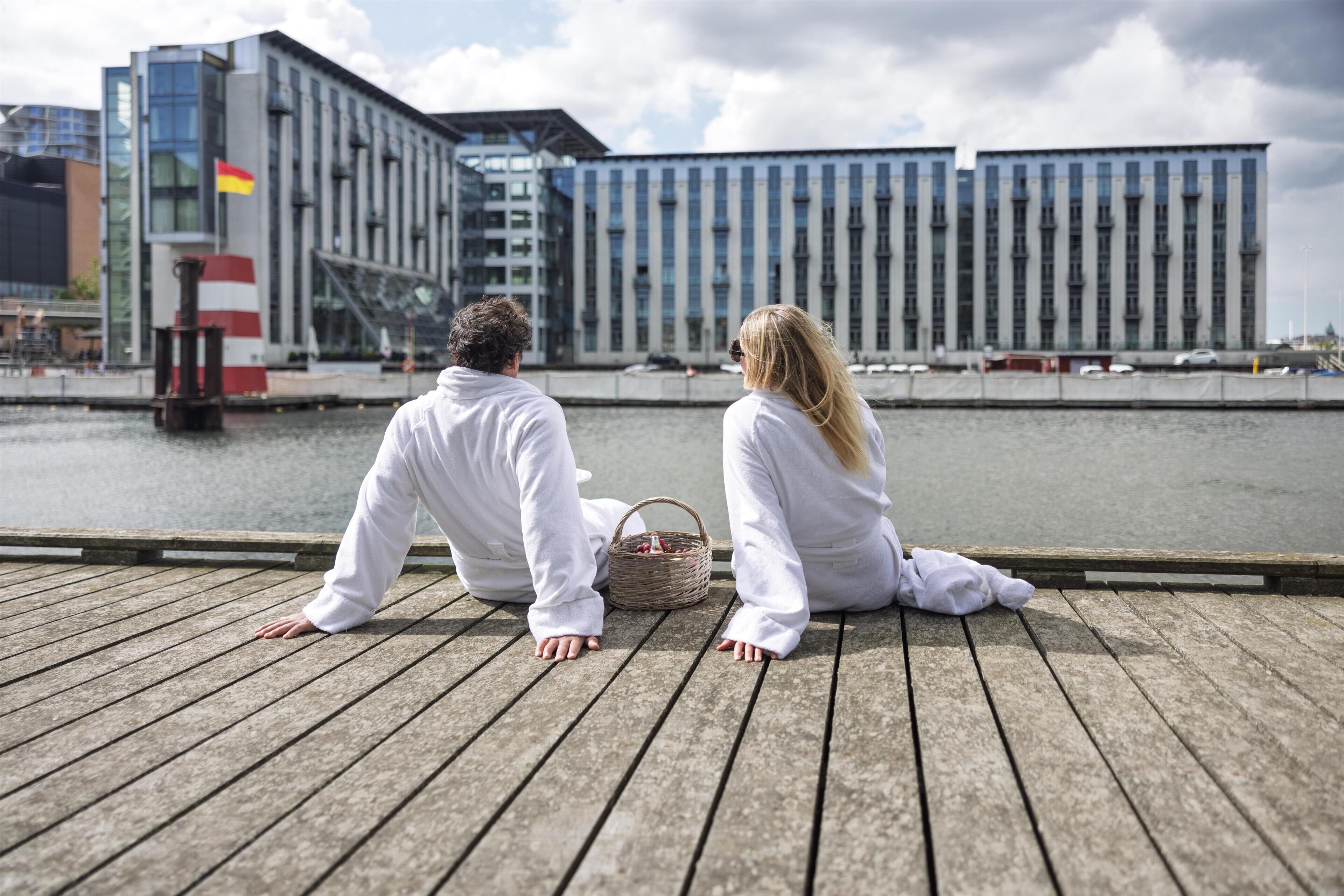 Two people sitting on a wooden dock overlooking a body of water with modern buildings in the background.
