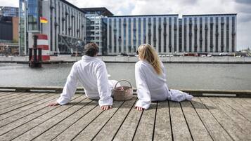 Two people sitting on a wooden dock overlooking a body of water with modern buildings in the background.