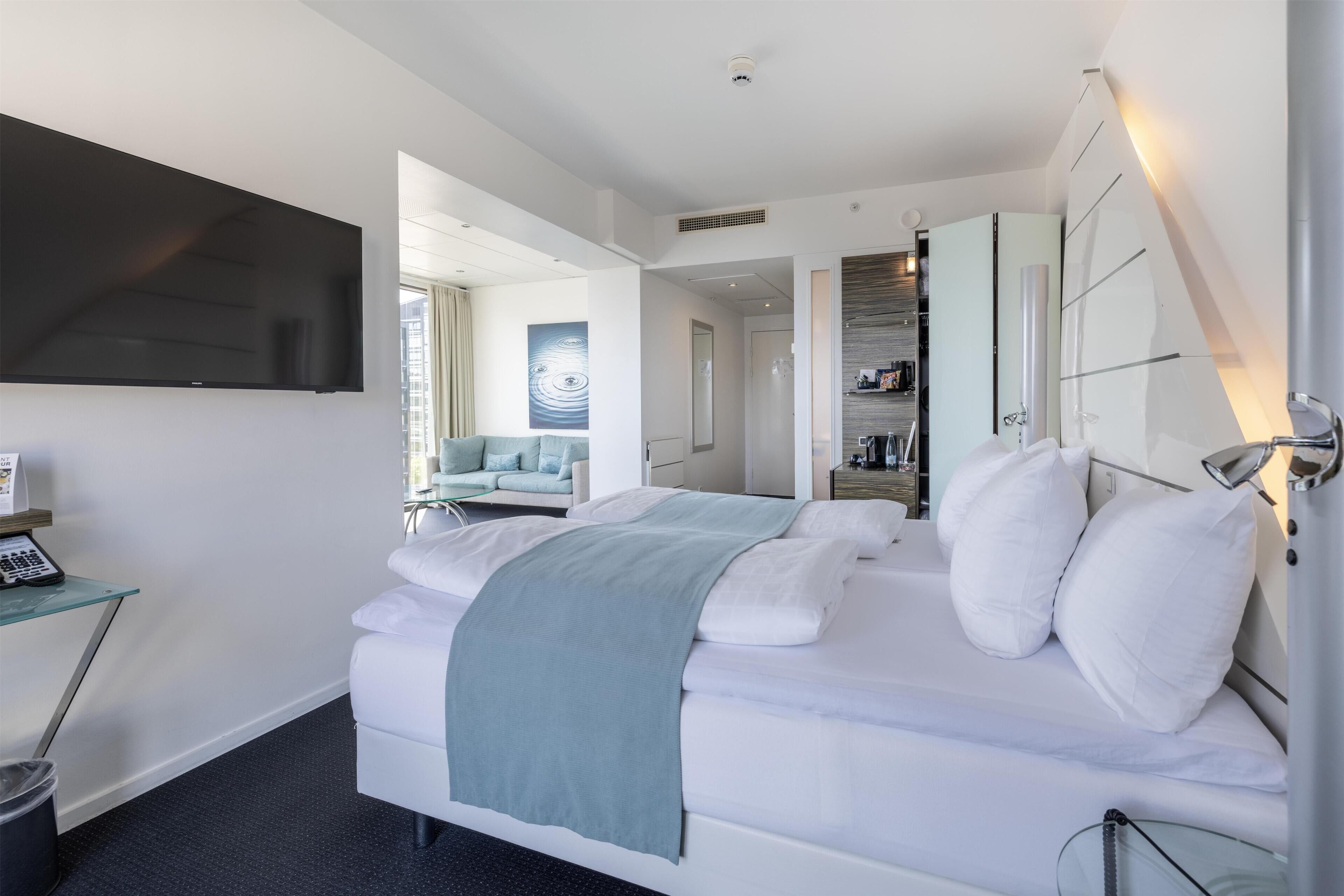 A modern hotel room with a large bed, a flat-screen TV, a desk with a telephone, and a bathroom with a mirror and towel rack.