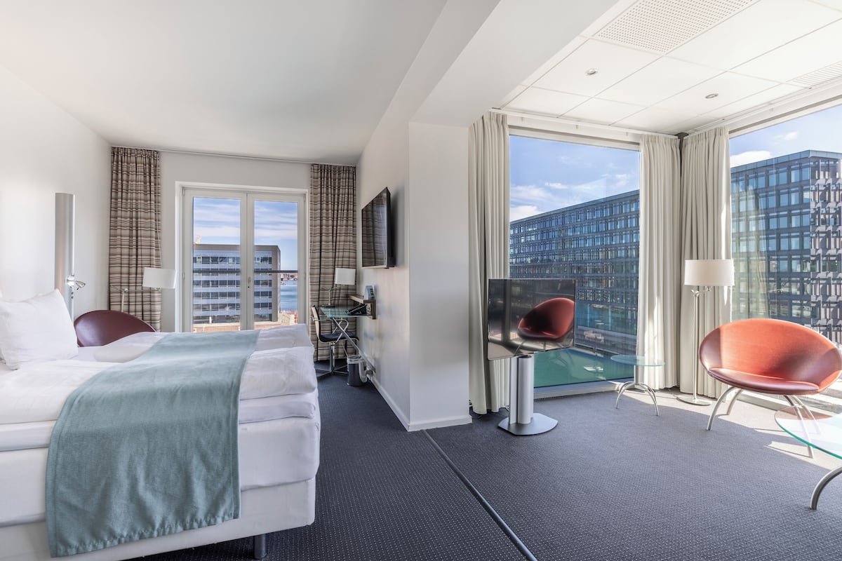 Une chambre d’hôtel moderne équipée d’un lit, d’un bureau, d’une chaise et d’une grande fenêtre donnant sur la ville.