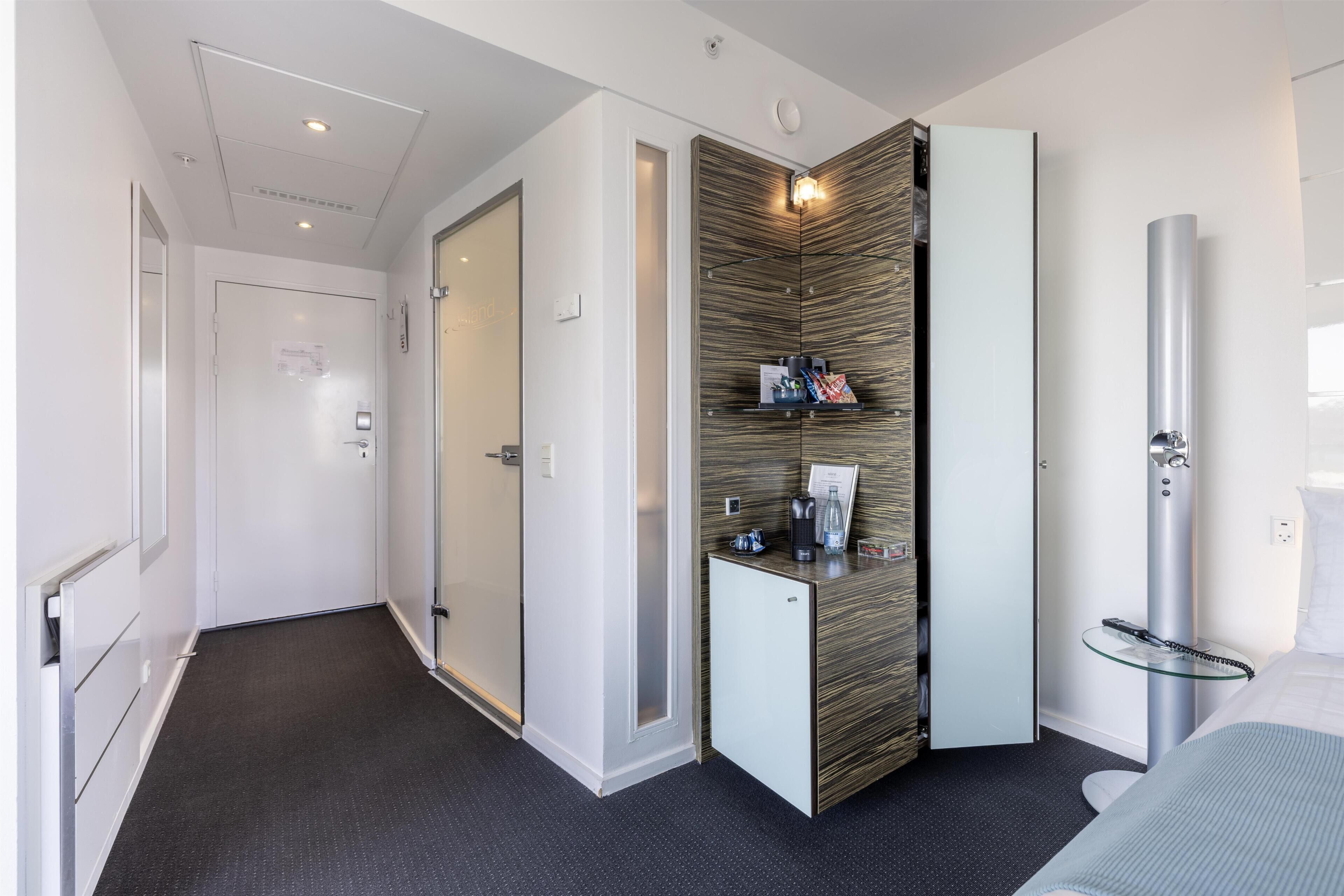 Habitación de hotel con diseño moderno, que presenta una alfombra oscura, un minibar, un espejo grande y una cama con cabecero blanco.
