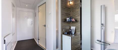 Habitación de hotel con diseño moderno, que presenta una alfombra oscura, un minibar, un espejo grande y una cama con cabecero blanco.