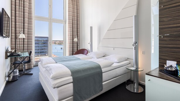 Habitación de hotel moderna con una cama grande, un escritorio con silla, una lámpara de escritorio y vistas de la ciudad a través de una ventana.
