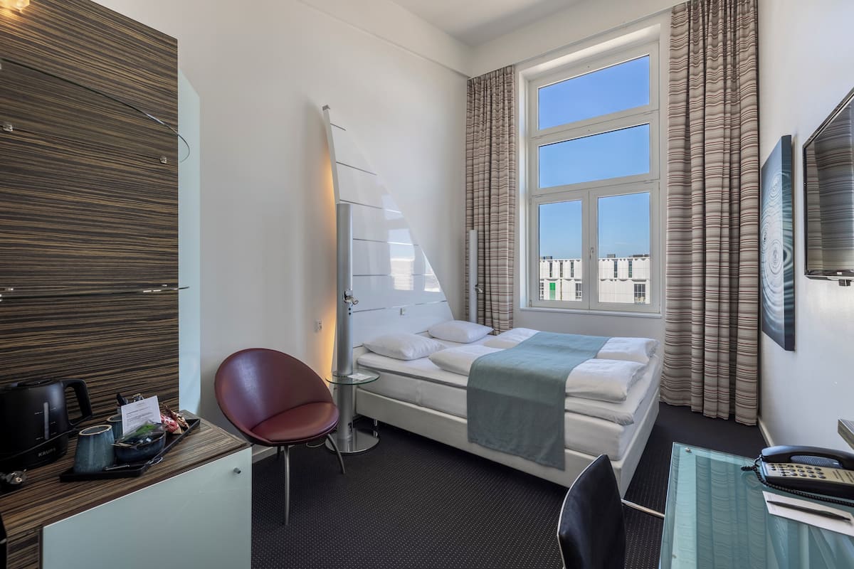 Une chambre d’hôtel comprenant un lit, un bureau avec un téléphone, une chaise, une lampe de bureau, une fenêtre avec des rideaux et une vue sur les bâtiments environnants.