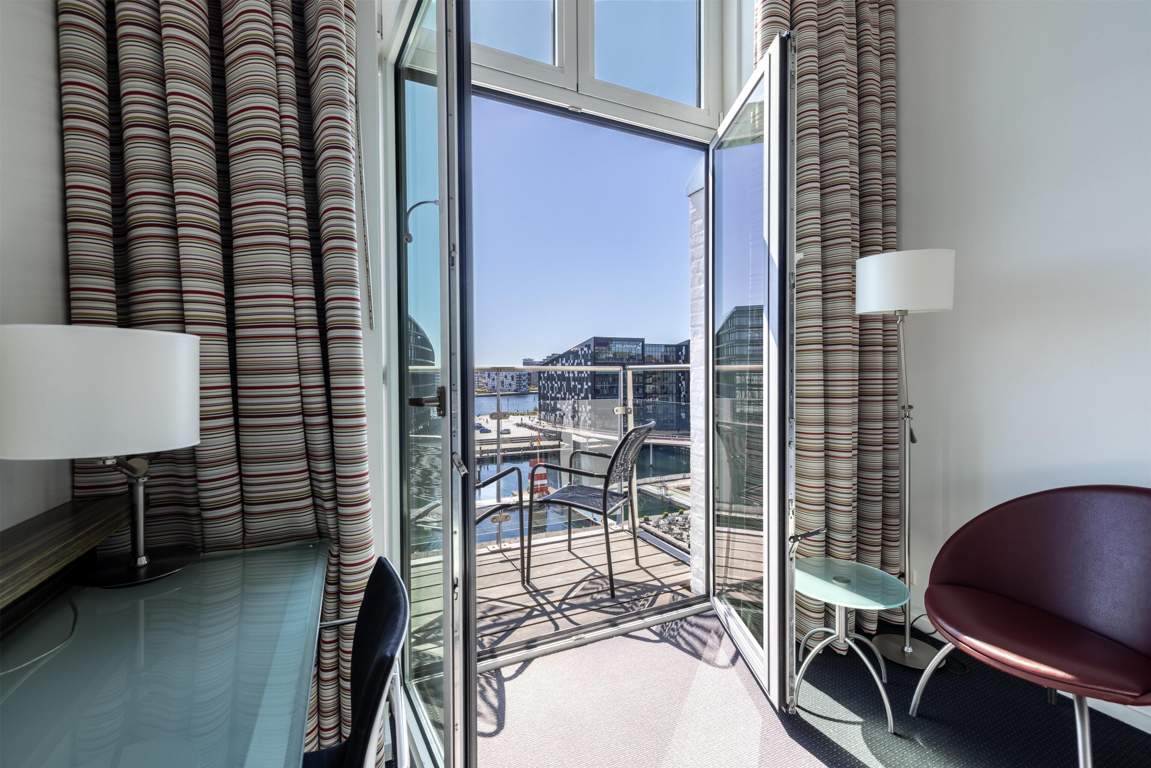 Habitación de hotel moderna con balcón, vista al puerto y zona de estar con mesa y sillas.