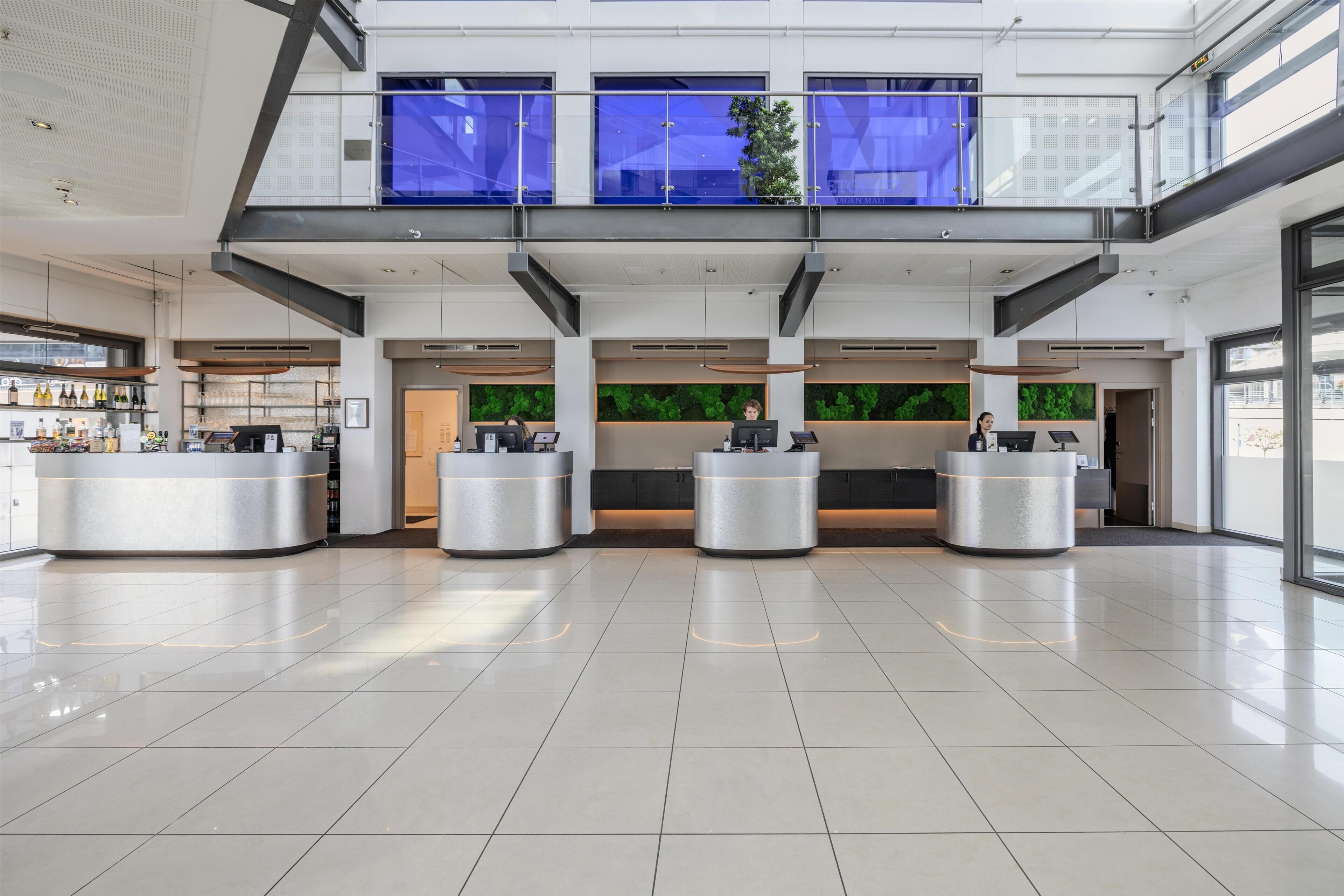 A spacious lobby with a modern design, featuring a central reception desk, a second-floor balcony with blue curtains, and a green wall with a plant.
