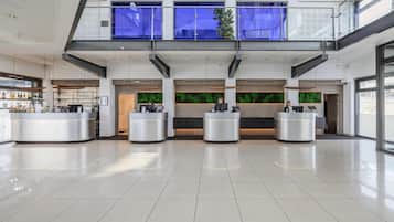 A spacious lobby with a modern design, featuring a central reception desk, a second-floor balcony with blue curtains, and a green wall with a plant.