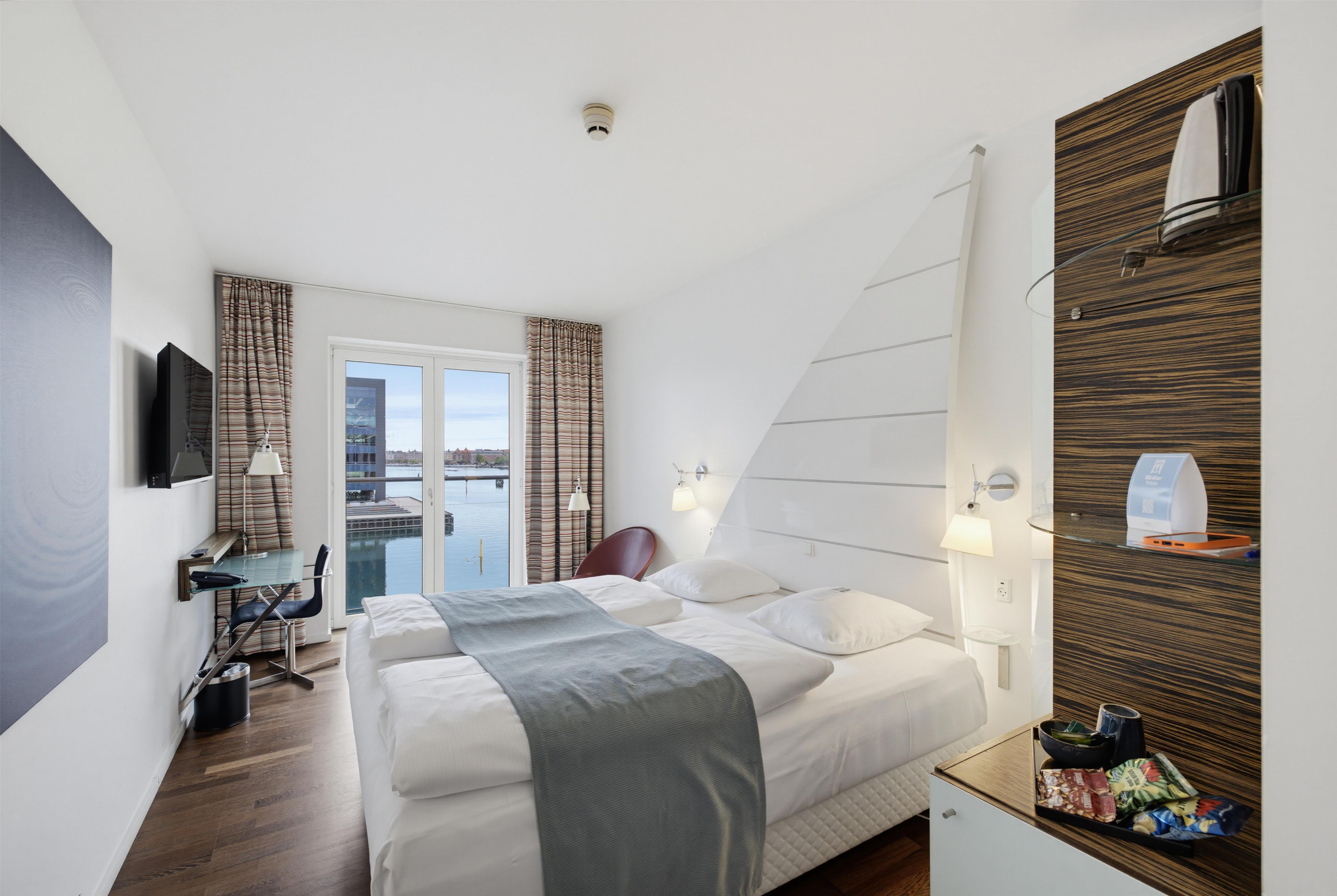 A hotel room with a bed, a desk, a chair, a TV, a window with curtains, and a view of the water.