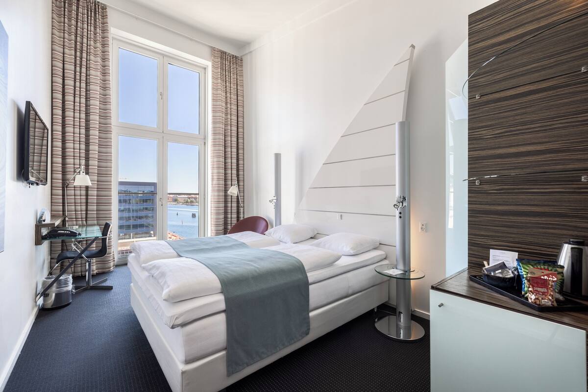 Une chambre d’hôtel moderne dotée d’un grand lit, d’un bureau avec une chaise, d’une petite table et offrant une vue sur la ville.