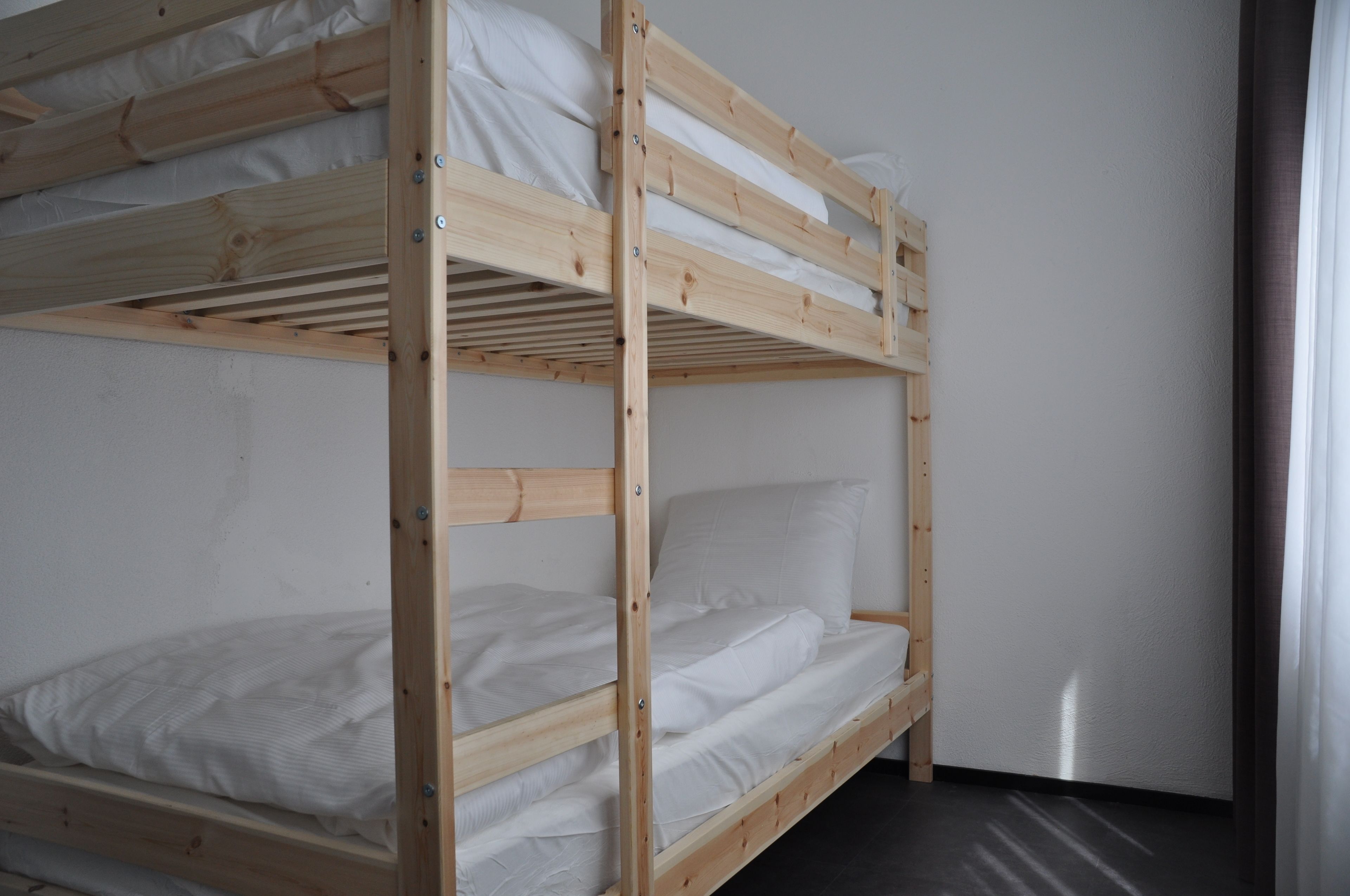 A bunk bed with white bedding in a room with plain walls and a window with curtains.