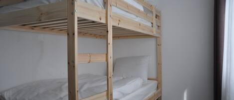 A bunk bed with white bedding in a room with plain walls and a window with curtains.