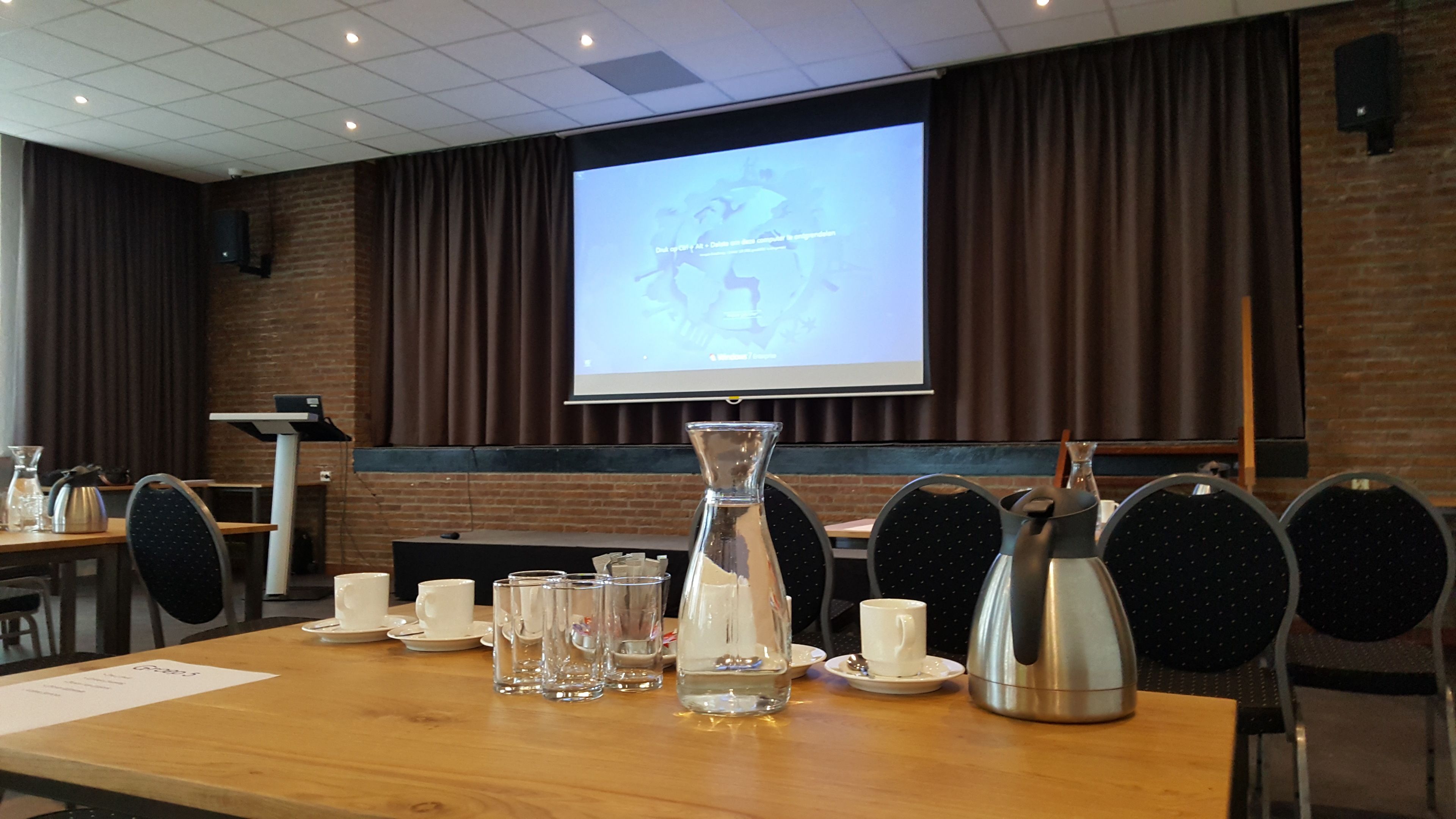 Un salón de conferencias con una pantalla grande mostrando una presentación, una mesa de madera con tazas y vasos, y una cafetera.