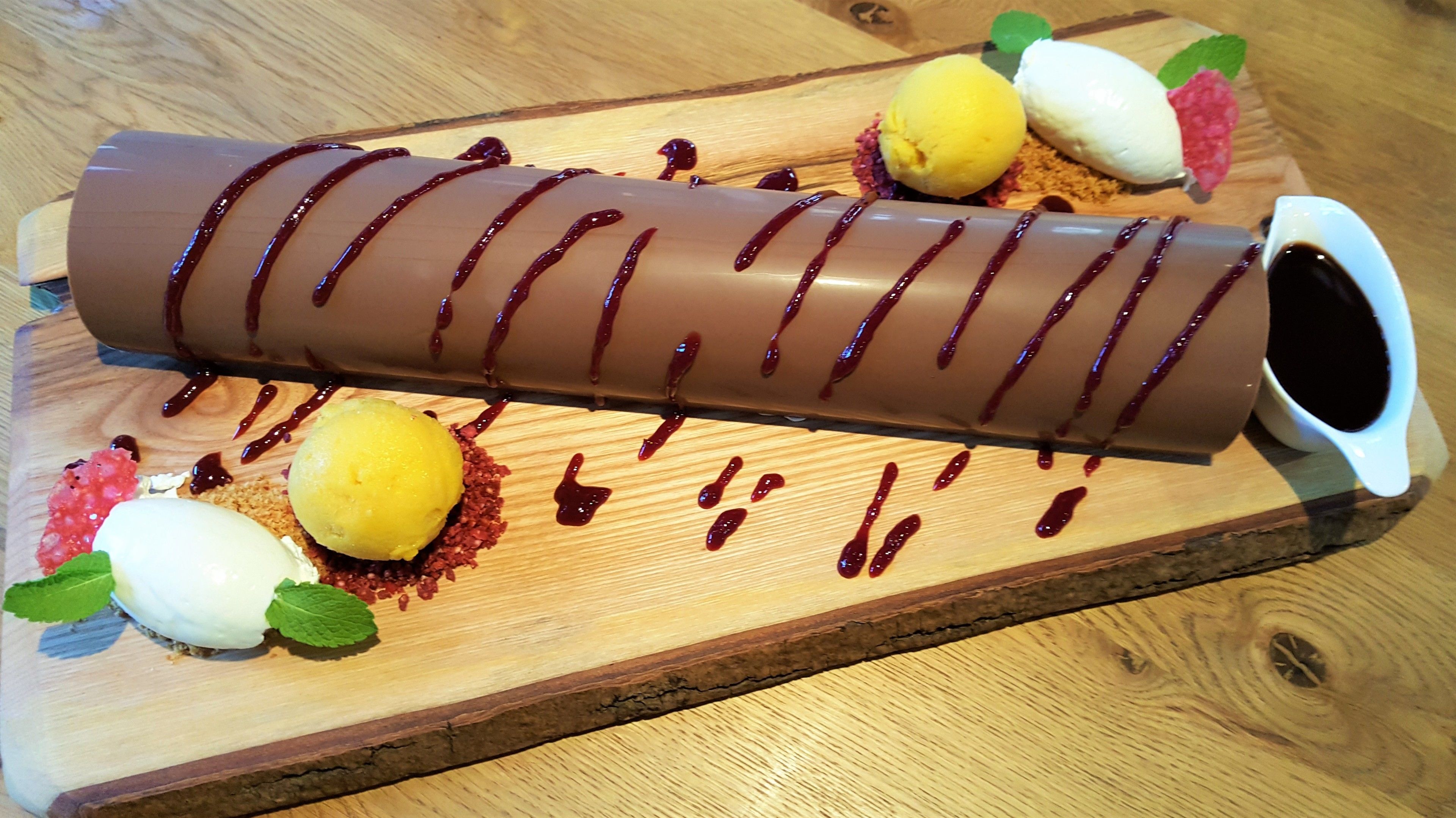 Postre cilíndric cobert de xocolata, amb escurrims, acompanyat d'una tassa blanca i dues rodanxes de llimona sobre una taula de fusta.