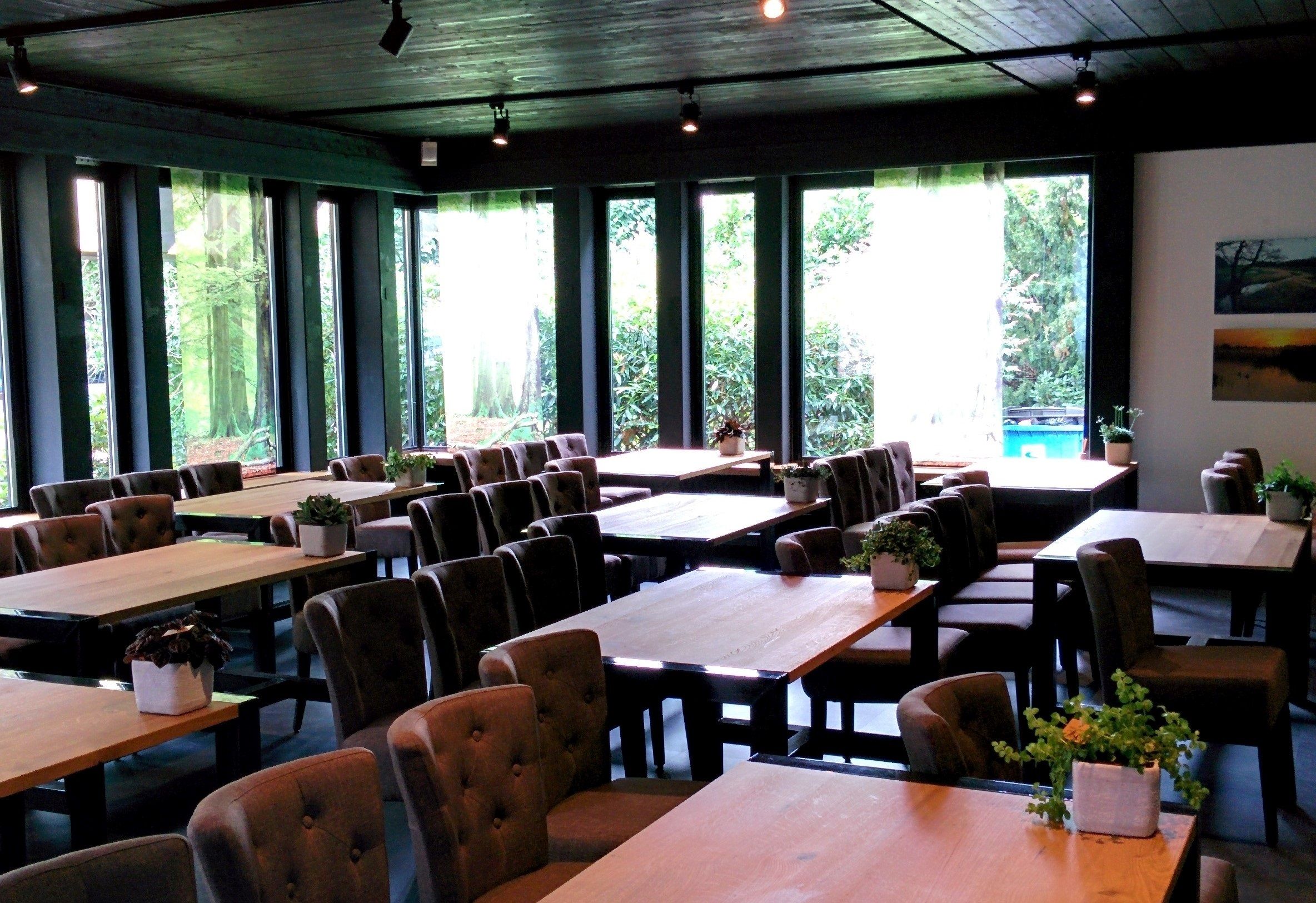 A restaurant with multiple tables and chairs, large windows with a view of greenery, and indoor lighting.