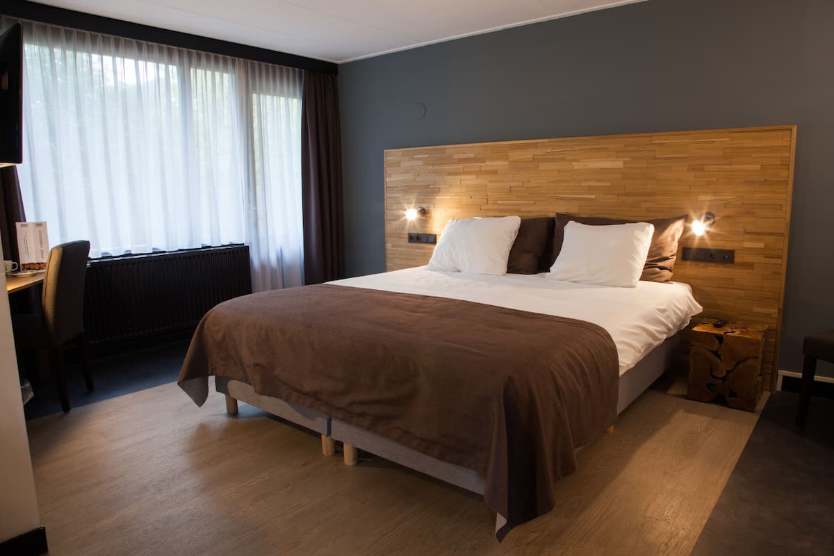 Hotellihuone, jossa on puinen sängynpääty, valkoisilla ja ruskeilla tekstiileillä varustettu sänky, työpöytä tuolilla sekä ikkuna, jossa on verhot.
