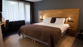 A hotel room with a wooden headboard, a bed with white and brown bedding, a desk with a chair, and a window with curtains.