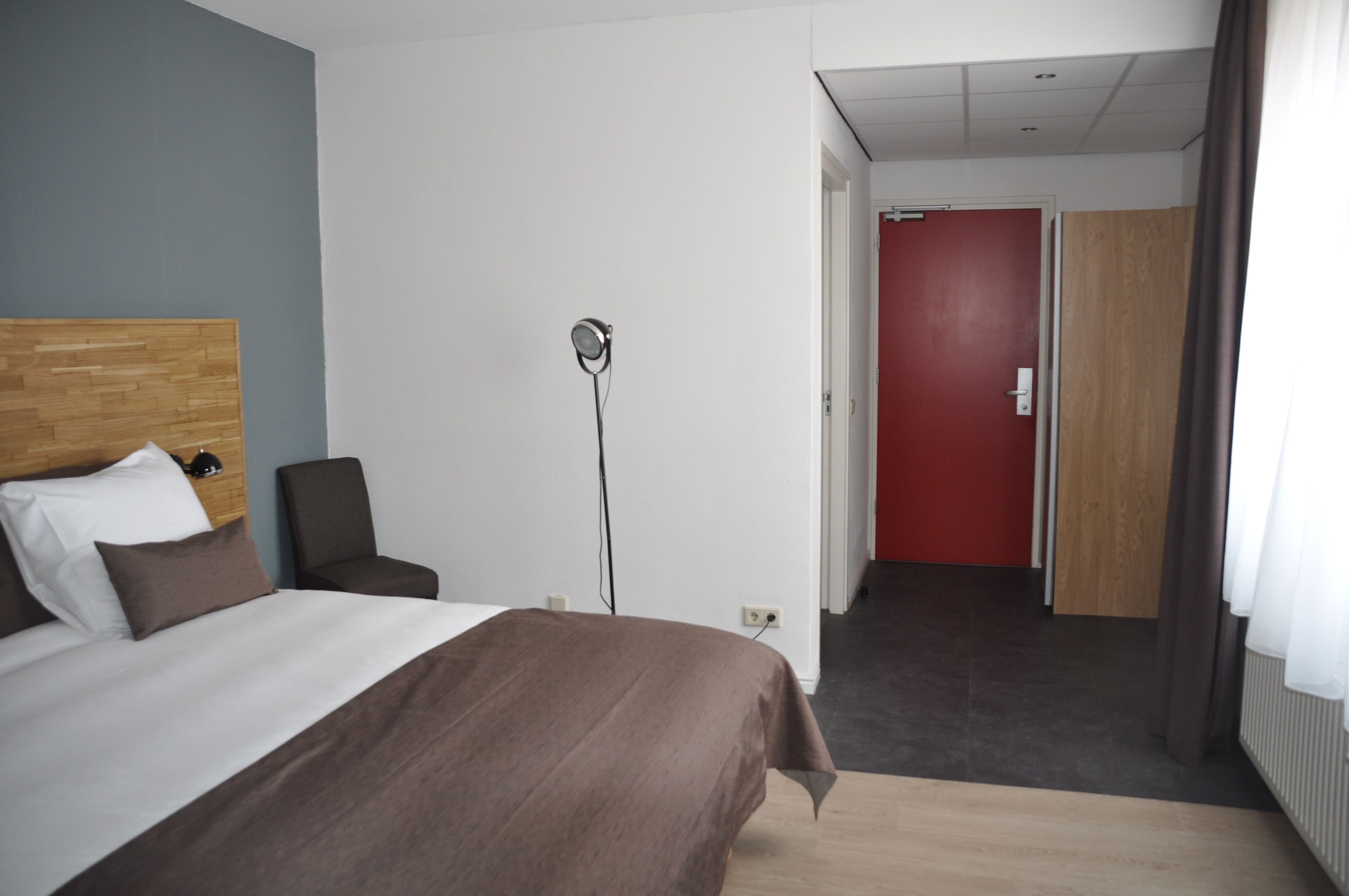 A hotel room with a bed, a bedside table, a lamp, and a view of a hallway with a red door.