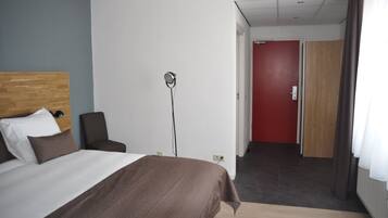 A hotel room with a bed, a bedside table, a lamp, and a view of a hallway with a red door.