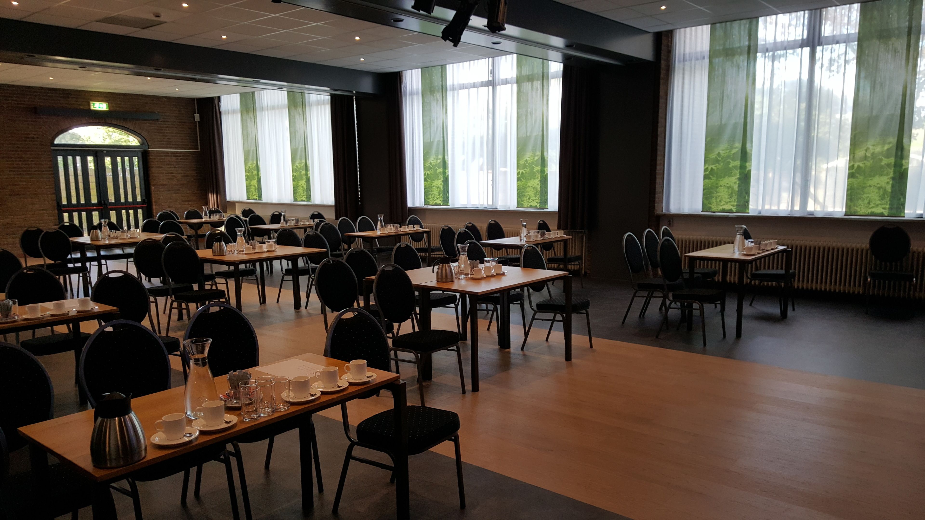 A conference room with multiple tables and chairs, large windows with curtains, and a brick wall with an emergency exit sign.