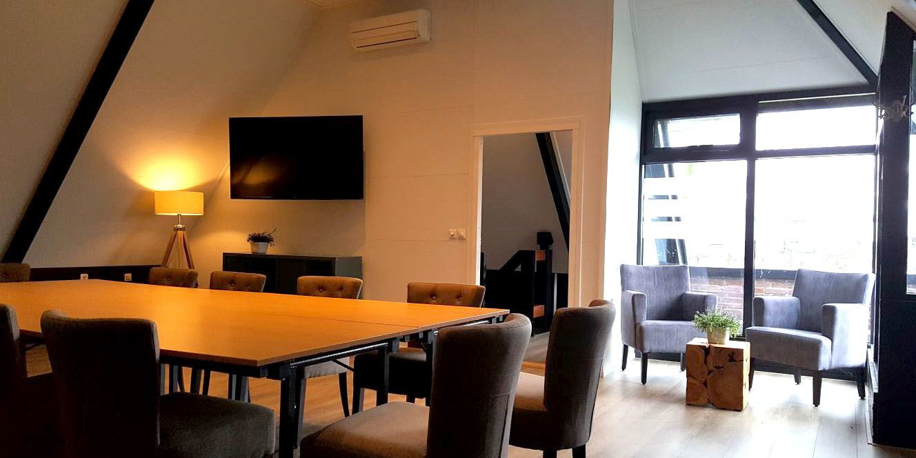 A modern conference room with a long table and comfortable chairs, a flat-screen TV, and a large window providing natural light.
