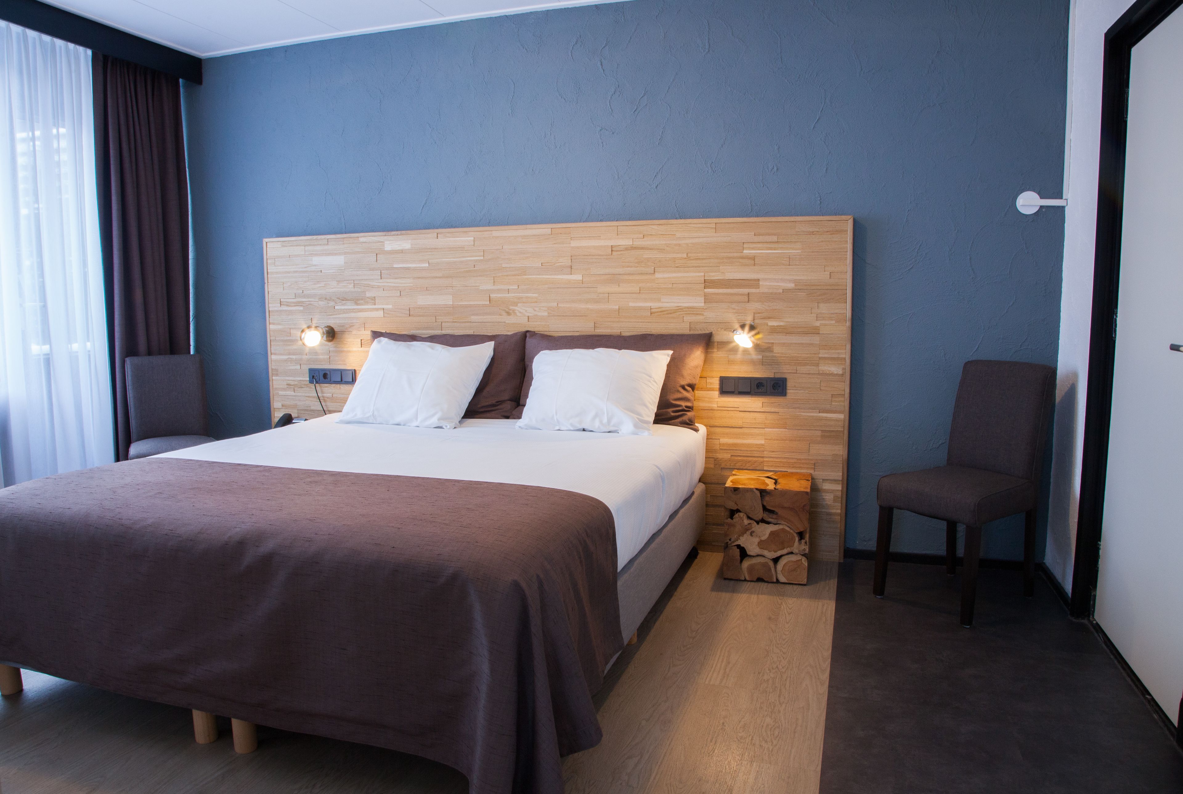 Een moderne hotelkamer met een houten hoofdeinde, een groot bed met een bruine sprei, twee nachtkastjes met lampen, een stoel en een klein houten bijzettafeltje.