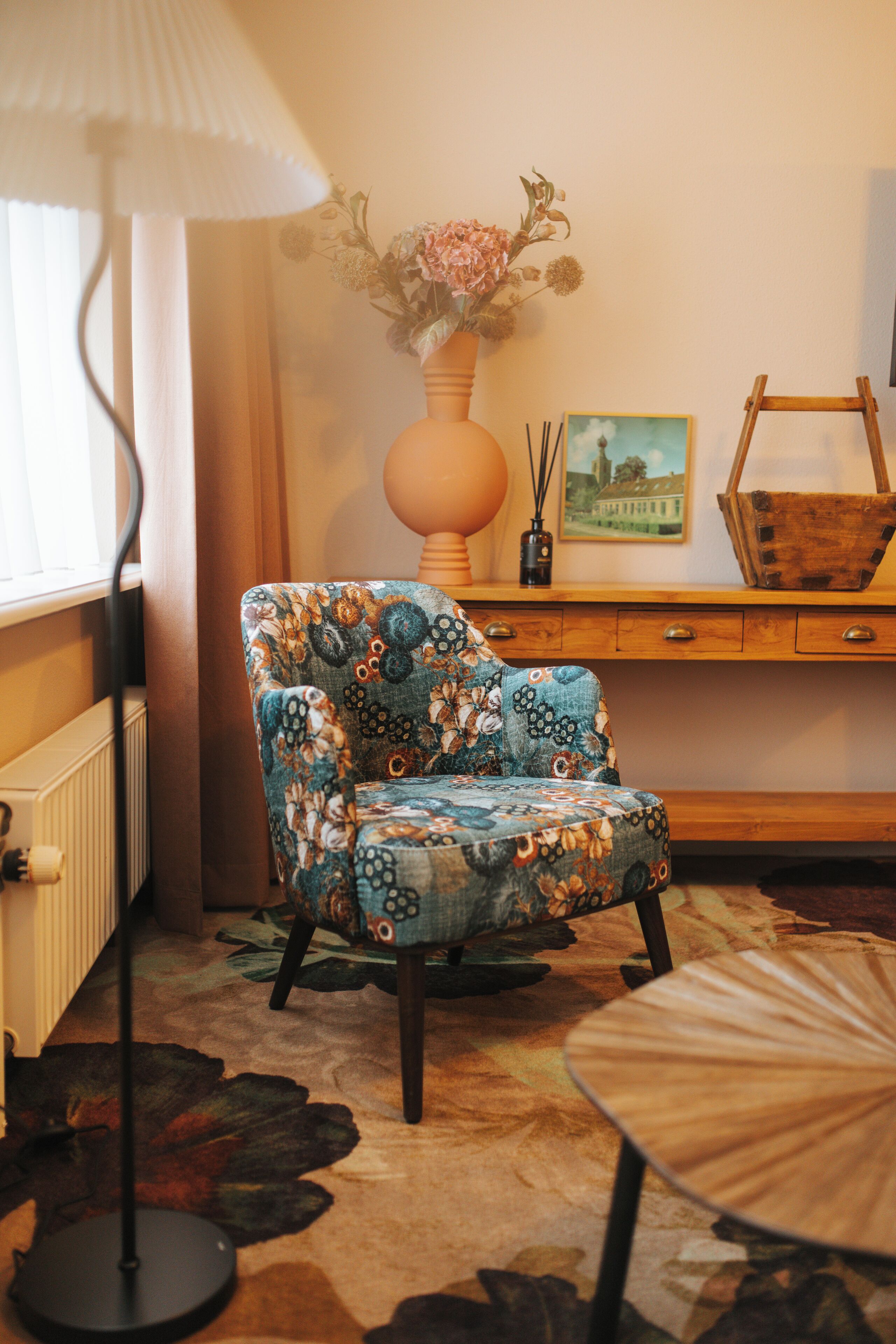 Ein Zimmer mit einem gemusterten Sessel, einer Lampe, einer Vase mit Blumen, einem gerahmten Bild und einem Holztisch.