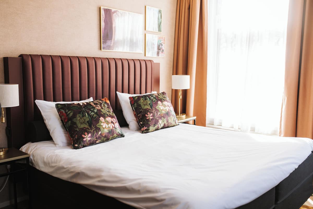 Habitación de hotel con una cama grande, dos mesitas de noche, una lámpara de pared y una ventana con cortinas.