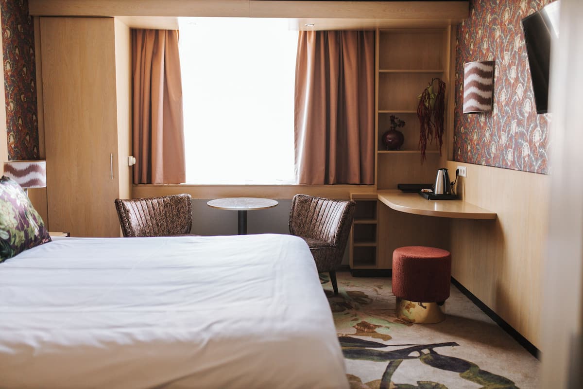 Habitación de hotel con cama, dos sillas, una mesita pequeña, una mesita auxiliar, una planta y una pared con diseño.