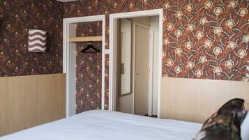 A hotel room with a patterned wallpaper, a bed with white bedding, a hanging garment, and a door leading to another room.