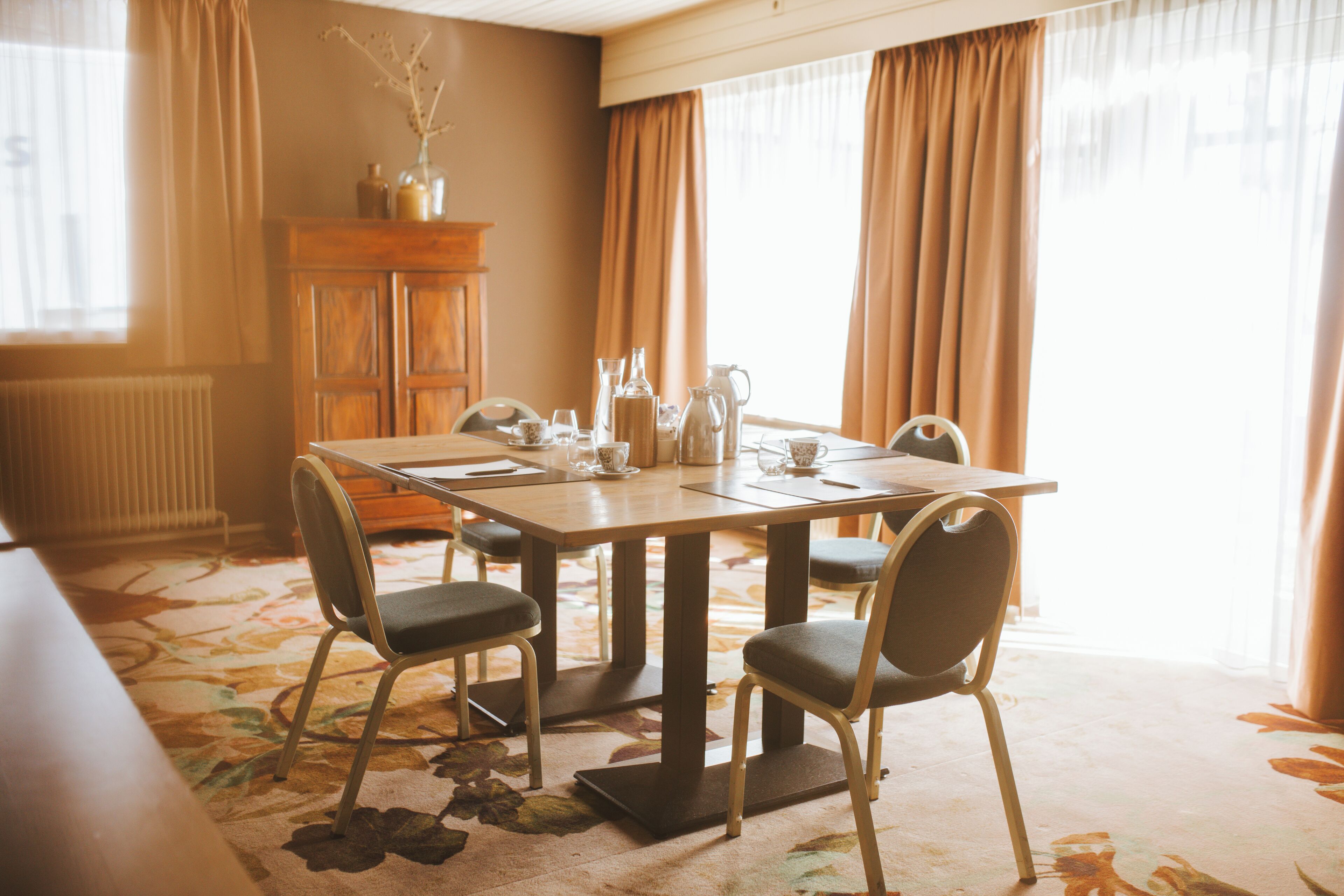 A dining area with a table set for four, surrounded by chairs. A wooden cabinet is against the wall, and a radiator is visible to the left.