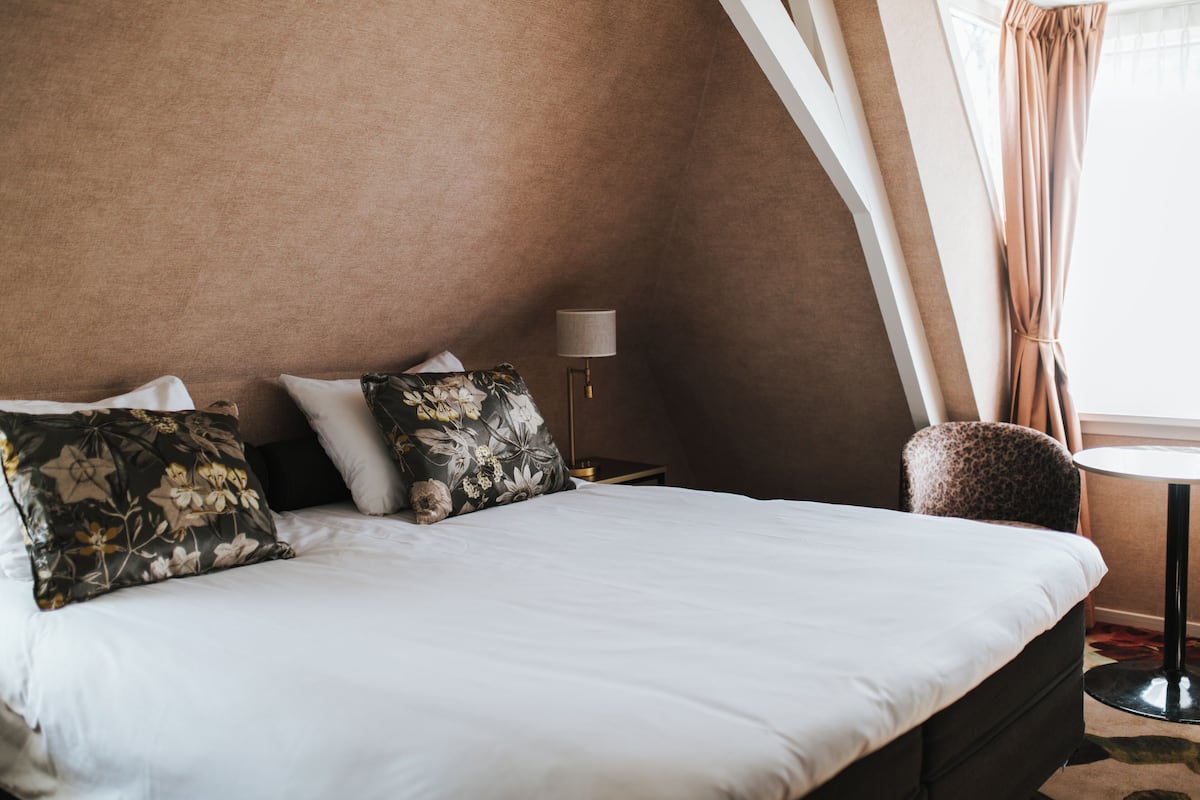 Una habitación de hotel con cama, mesita de noche, lámpara, una silla y ventana con cortinas.