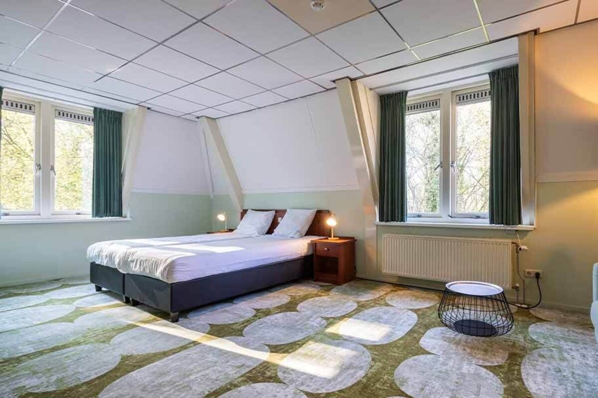 A bedroom with a bed, bedside table, lamp, and a view of the outdoors through a window.