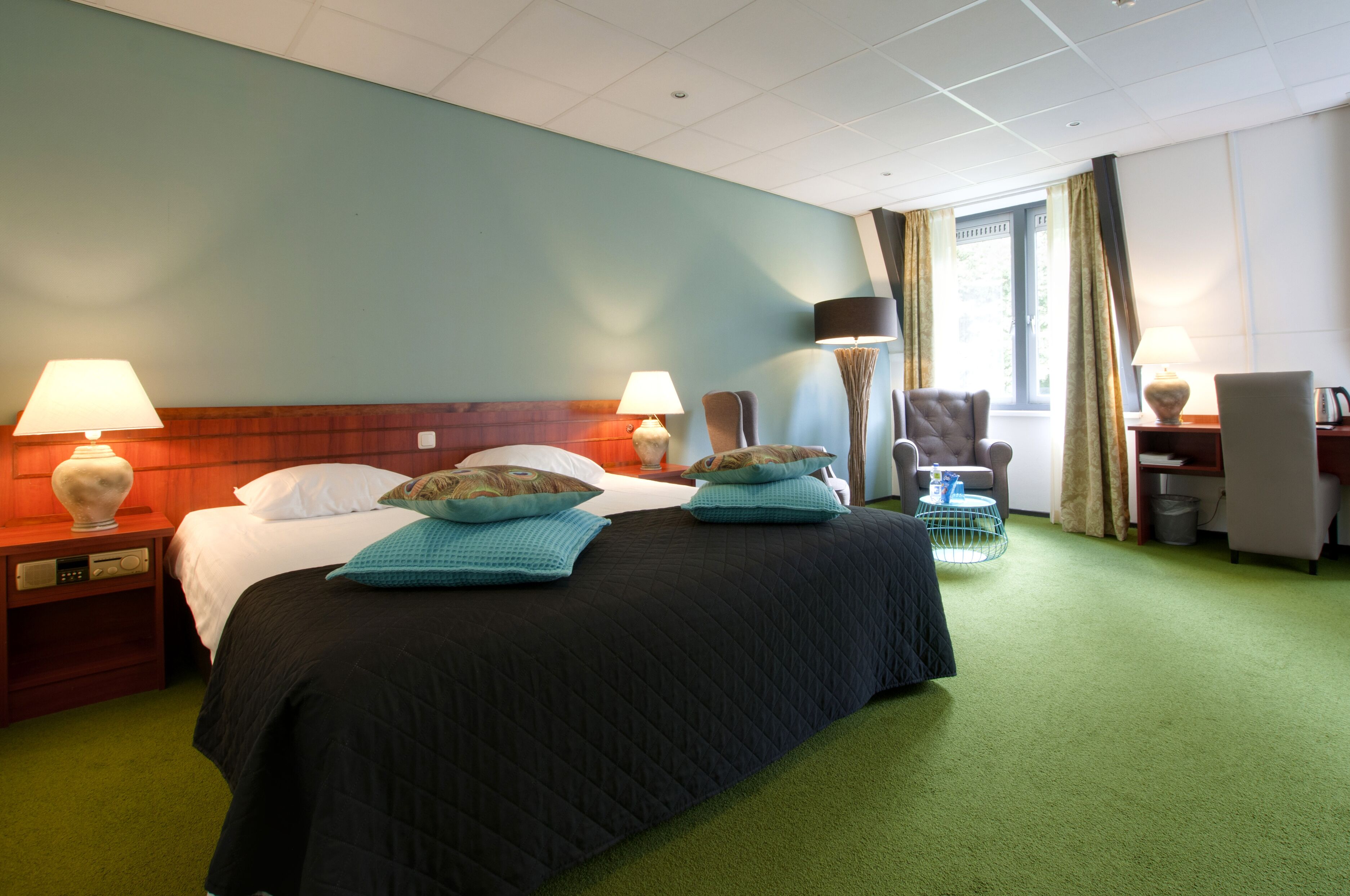A hotel room with a large bed, two bedside lamps, a desk with a chair, and a window with curtains.