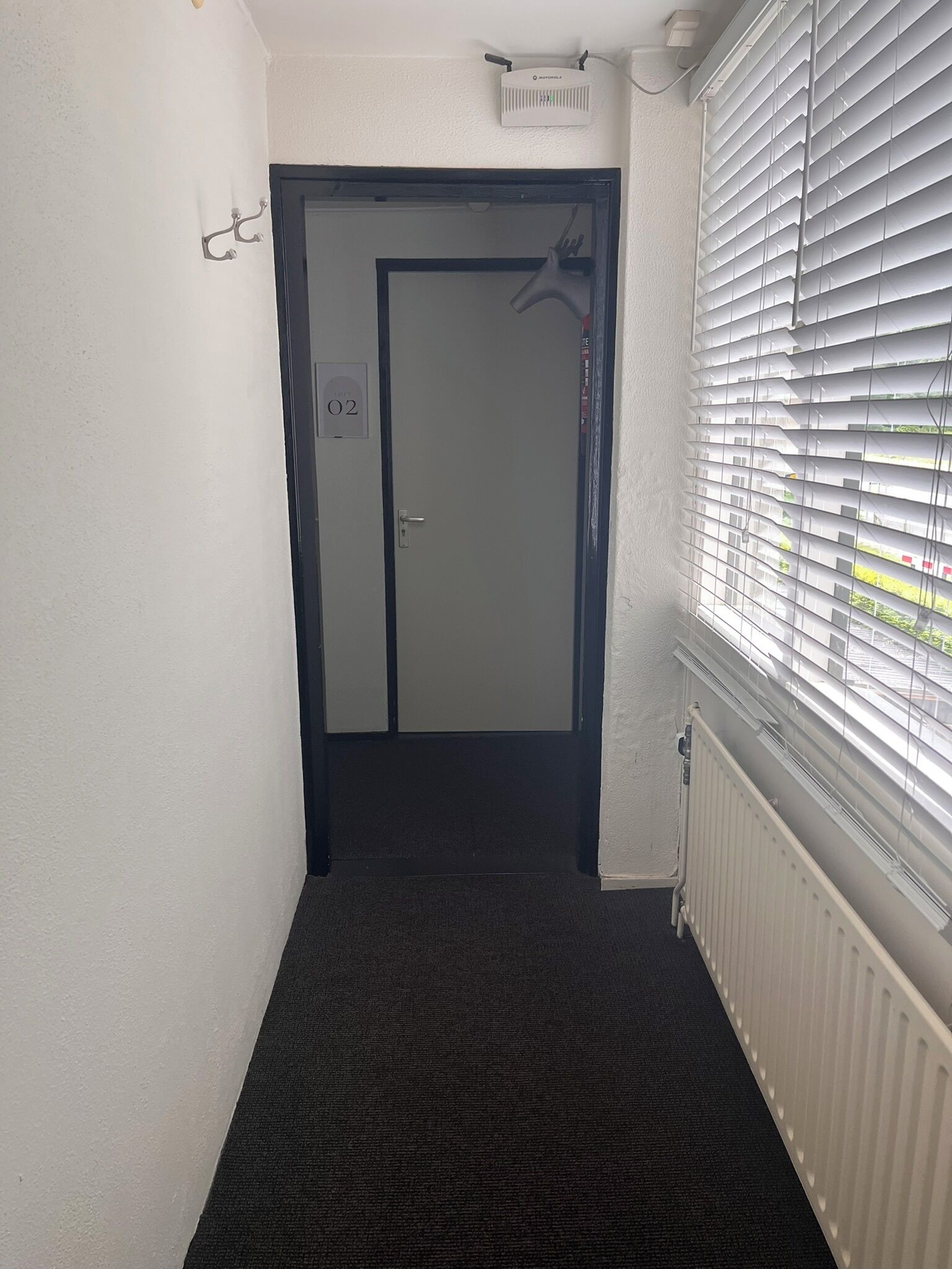 Ein schmaler Korridor mit Teppichboden, einer Tür mit der Aufschrift „02“ und einem Heizkörper an der rechten Seite.