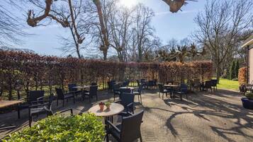Een terras buiten met tafels en stoelen, omringd door heggen en bomen.